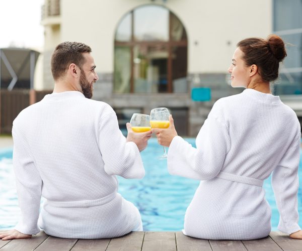 Idyllic couple toasting with glasses of juice while sitting by swimming-pool